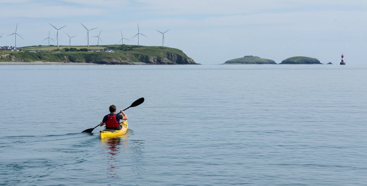 A solo paddler in a yellow sit-on-top kayak and red buoyancy aid mid-stroke on the calm blue-grey water of Skerries Bay in north County Dublin, the low offshore islets of St Patrick's Island and Colt Island visible on the horizon and the Skerries village windmills standing on the headland behind